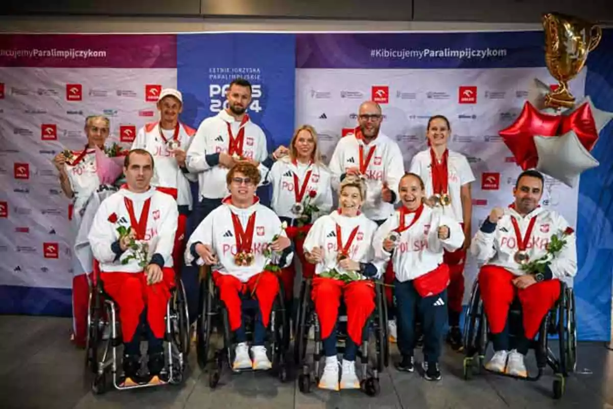 Un grupo de atletas paralímpicos polacos posan con medallas y flores frente a un fondo con logotipos y globos.