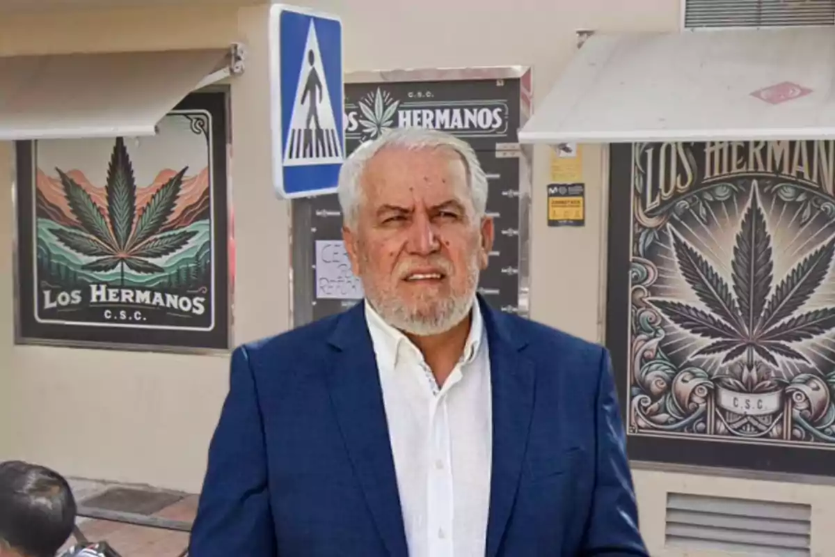 Hombre de cabello canoso y barba con traje azul y camisa blanca de pie frente a un local con carteles de hojas de marihuana y el nombre Los Hermanos