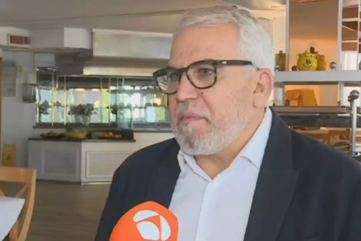 Hombre de cabello canoso y barba con gafas oscuras siendo entrevistado en un restaurante mientras sostiene un micrófono naranja Hombre de cabello canoso y barba con gafas oscuras siendo entrevistado en un restaurante mientras sostiene un micrófono naranja
