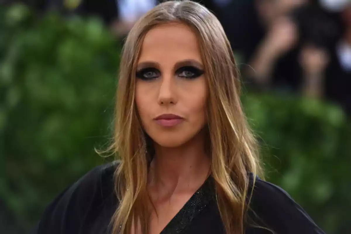 Una persona con cabello largo y rubio, maquillaje de ojos oscuro y labios rosados, vistiendo una prenda negra, con un fondo desenfocado de vegetación. Una persona con cabello largo y rubio, maquillaje de ojos oscuro y labios rosados, vistiendo una prenda negra, con un fondo desenfocado de vegetación.