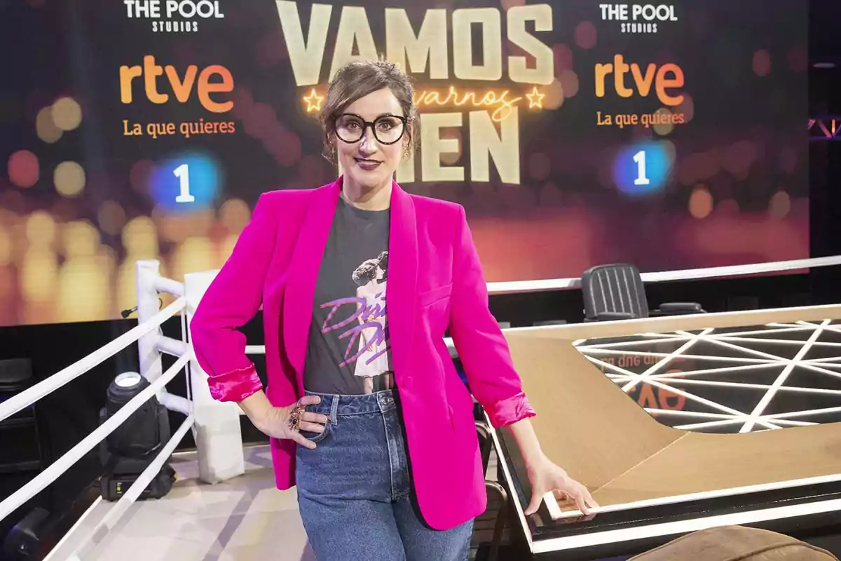 Mujer con gafas y chaqueta fucsia posando en un set de televisión con fondo que dice Vamos a llevarnos bien y logotipos de RTVE Mujer con gafas y chaqueta fucsia posando en un set de televisión con fondo que dice Vamos a llevarnos bien y logotipos de RTVE