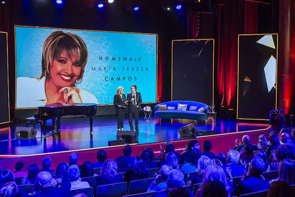 En la imagen se observa un escenario con una gran pantalla que muestra una fotografía de una mujer sonriente y el texto "Homenaje María Teresa Campos", mientras dos personas están de pie en el escenario frente a un piano de cola y una audiencia sentada.
