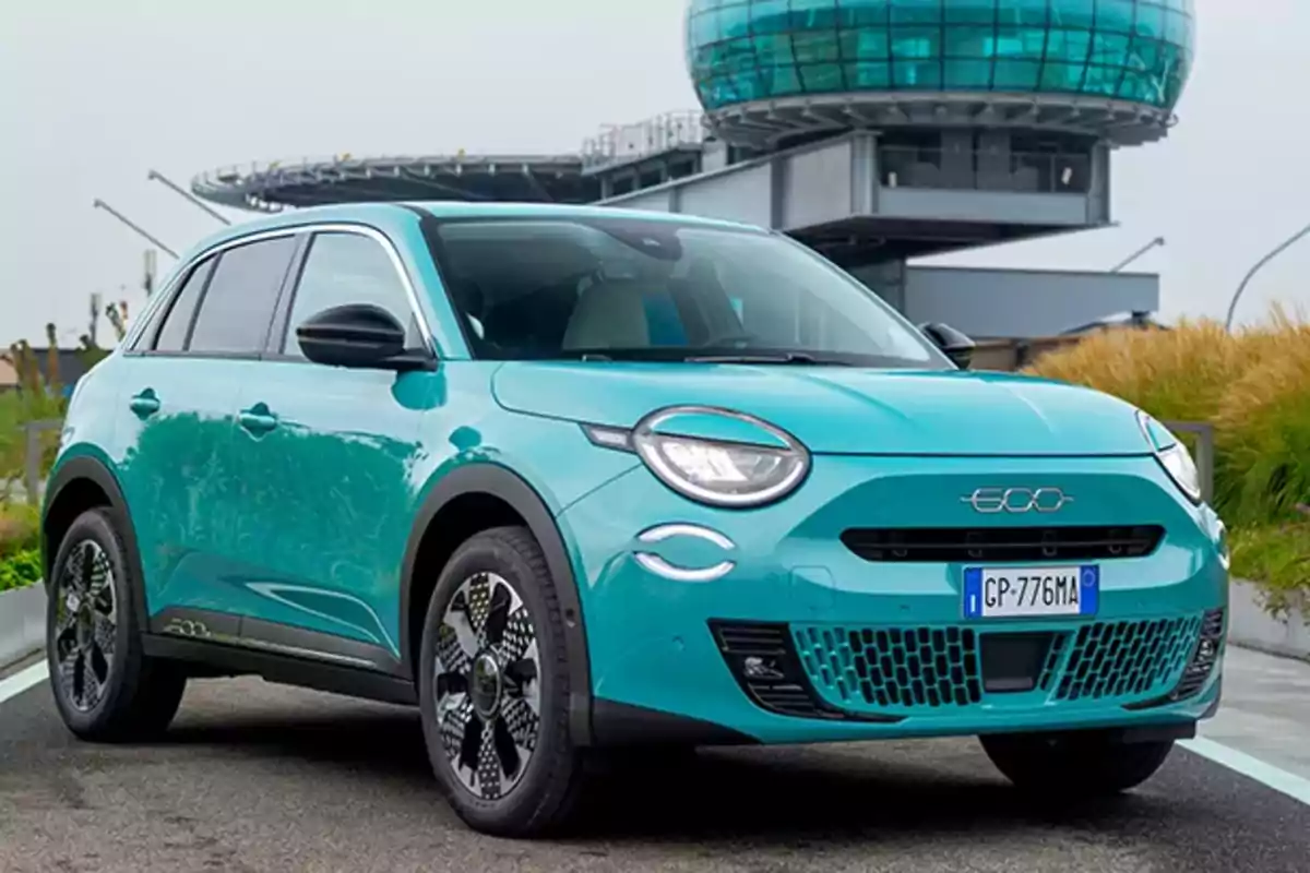 Un FIAT 600 Hybrid compacto azul turquesa estacionado en una calle con un edificio moderno al fondo.