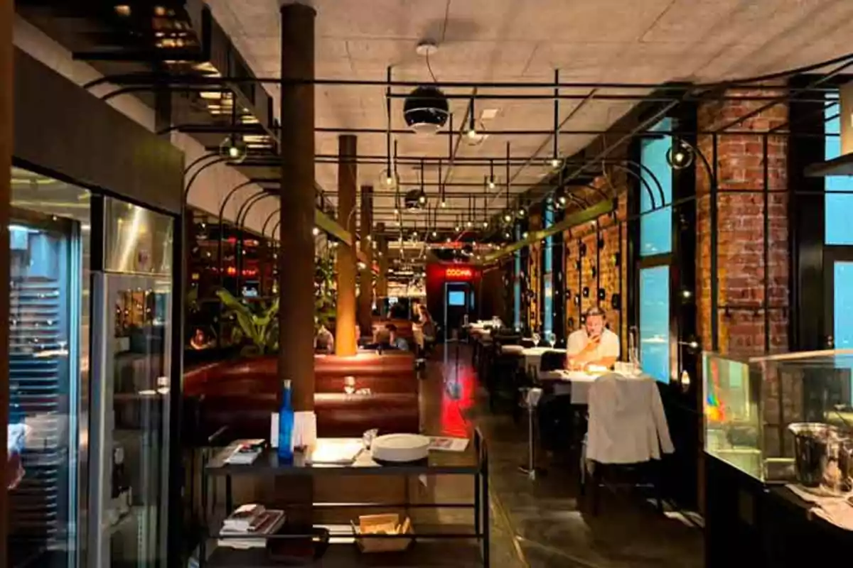 Interior de un restaurante moderno con mesas, sillas y decoración industrial, donde se observa a varias personas cenando.