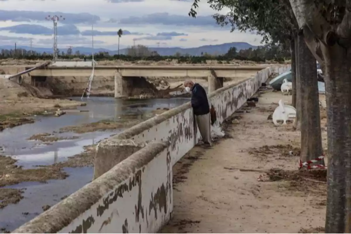 Un hombre con mascarilla se inclina sobre una barandilla junto a un río con un puente al fondo y árboles a la derecha.