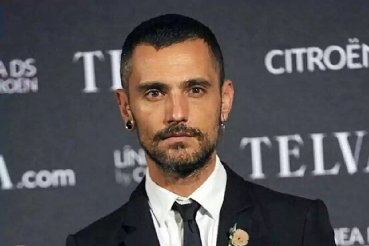 Hombre con barba y cabello corto vestido con traje negro y corbata posando frente a un fondo con logotipos de marcas Hombre con barba y cabello corto vestido con traje negro y corbata posando frente a un fondo con logotipos de marcas