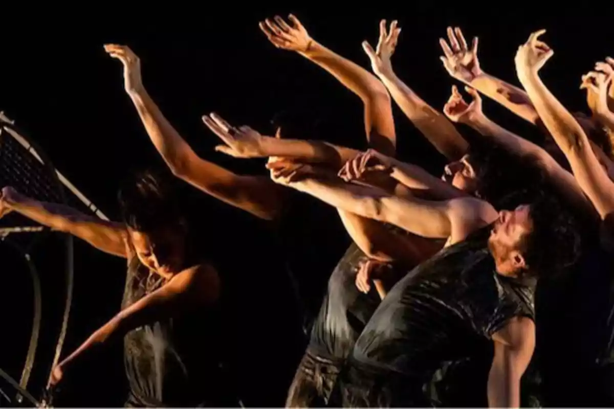 Grupo de bailarines en una coreografía contemporánea con movimientos expresivos y brazos extendidos.