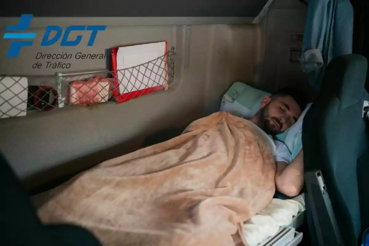 Un hombre durmiendo en la cabina de un camión con una manta beige, con el logo de la Dirección General de Tráfico (DGT) en la esquina superior izquierda.