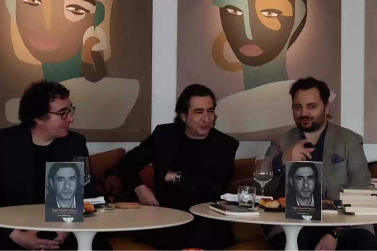 Tres hombres sentados en una mesa redonda con libros y bebidas, con dos cuadros abstractos en la pared detrás de ellos.