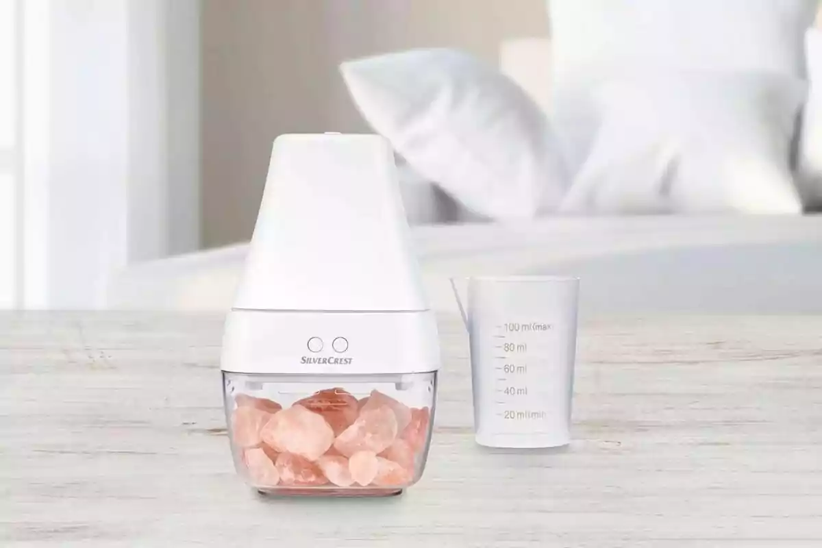 Un difusor blanco de Lidl con piedras rosadas en su base junto a un vaso medidor transparente sobre una superficie de madera clara en un entorno de dormitorio. Un difusor blanco de Lidl con piedras rosadas en su base junto a un vaso medidor transparente sobre una superficie de madera clara en un entorno de dormitorio.