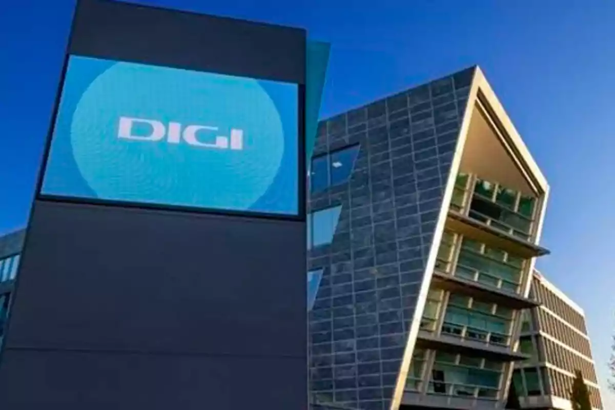 Edificio moderno con fachada de vidrio y pantalla que muestra el logo de DIGI bajo un cielo azul.