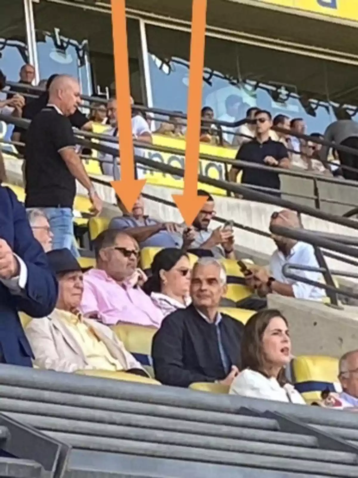 Personas sentadas en las gradas de un estadio, con dos flechas naranjas señalando a dos individuos específicos.