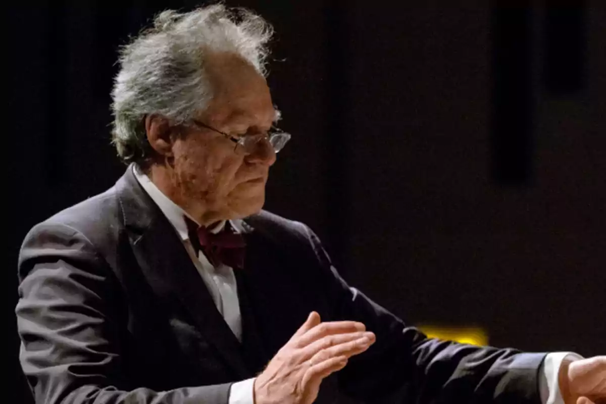 Un hombre mayor dirigiendo una orquesta con un traje formal y pajarita.