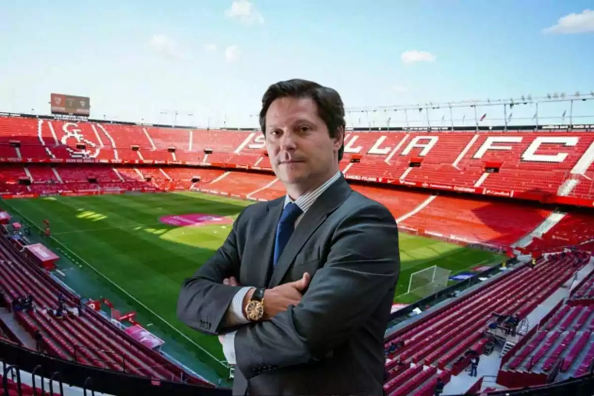 Hombre de traje con los brazos cruzados posando en un estadio de fútbol vacío con gradas rojas y el nombre Sevilla FC en el fondo