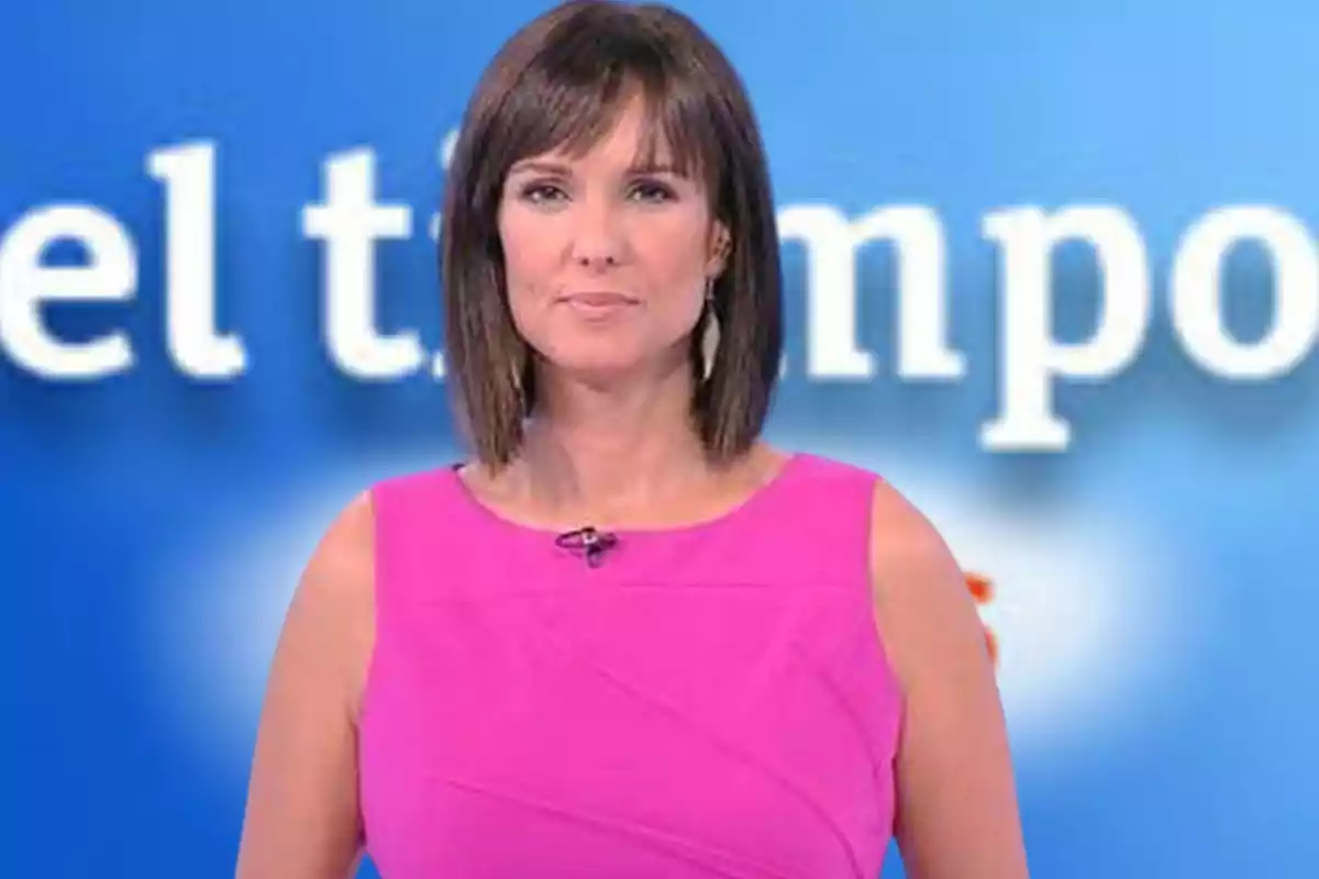 Presentadora de televisión con vestido rosa frente a un fondo azul con letras blancas desenfocadas