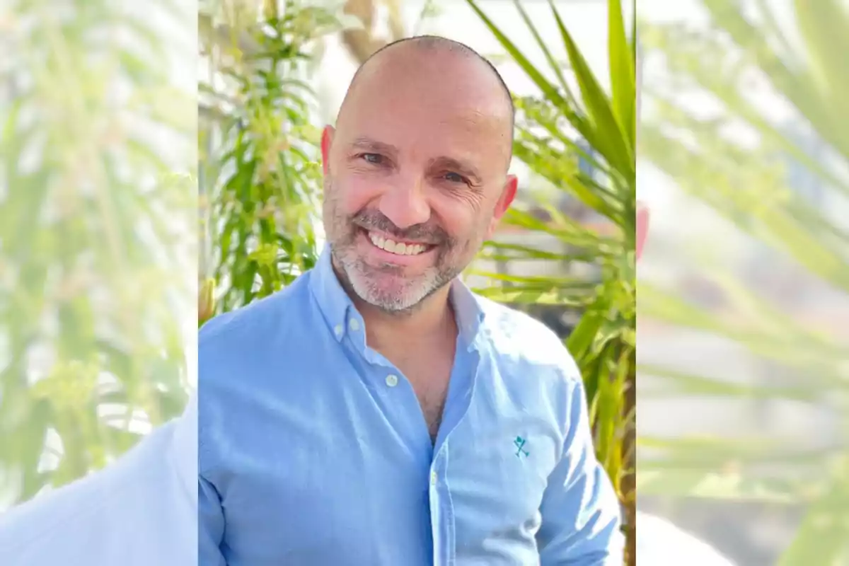 Hombre calvo con barba corta y camisa azul claro sonriendo frente a plantas verdes en un entorno soleado Hombre calvo con barba corta y camisa azul claro sonriendo frente a plantas verdes en un entorno soleado
