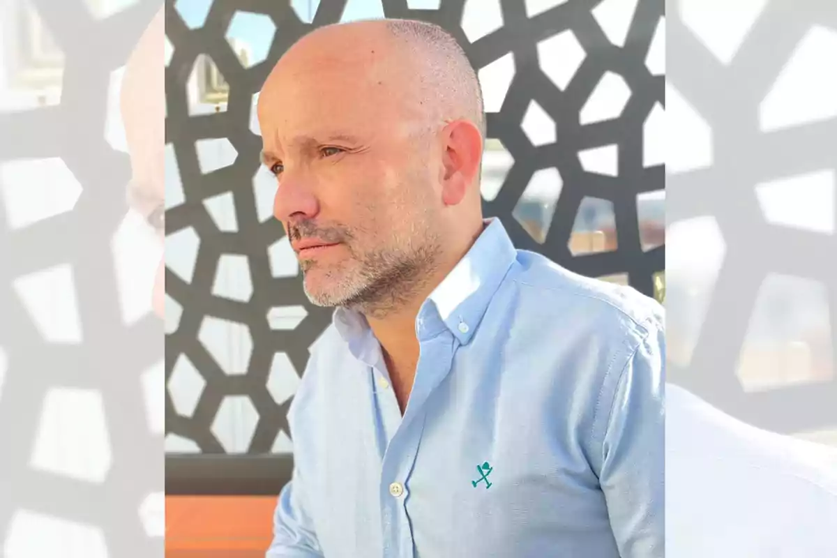 Hombre calvo con barba corta y camisa azul claro sentado frente a un fondo con patrón geométrico oscuro Hombre calvo con barba corta y camisa azul claro sentado frente a un fondo con patrón geométrico oscuro