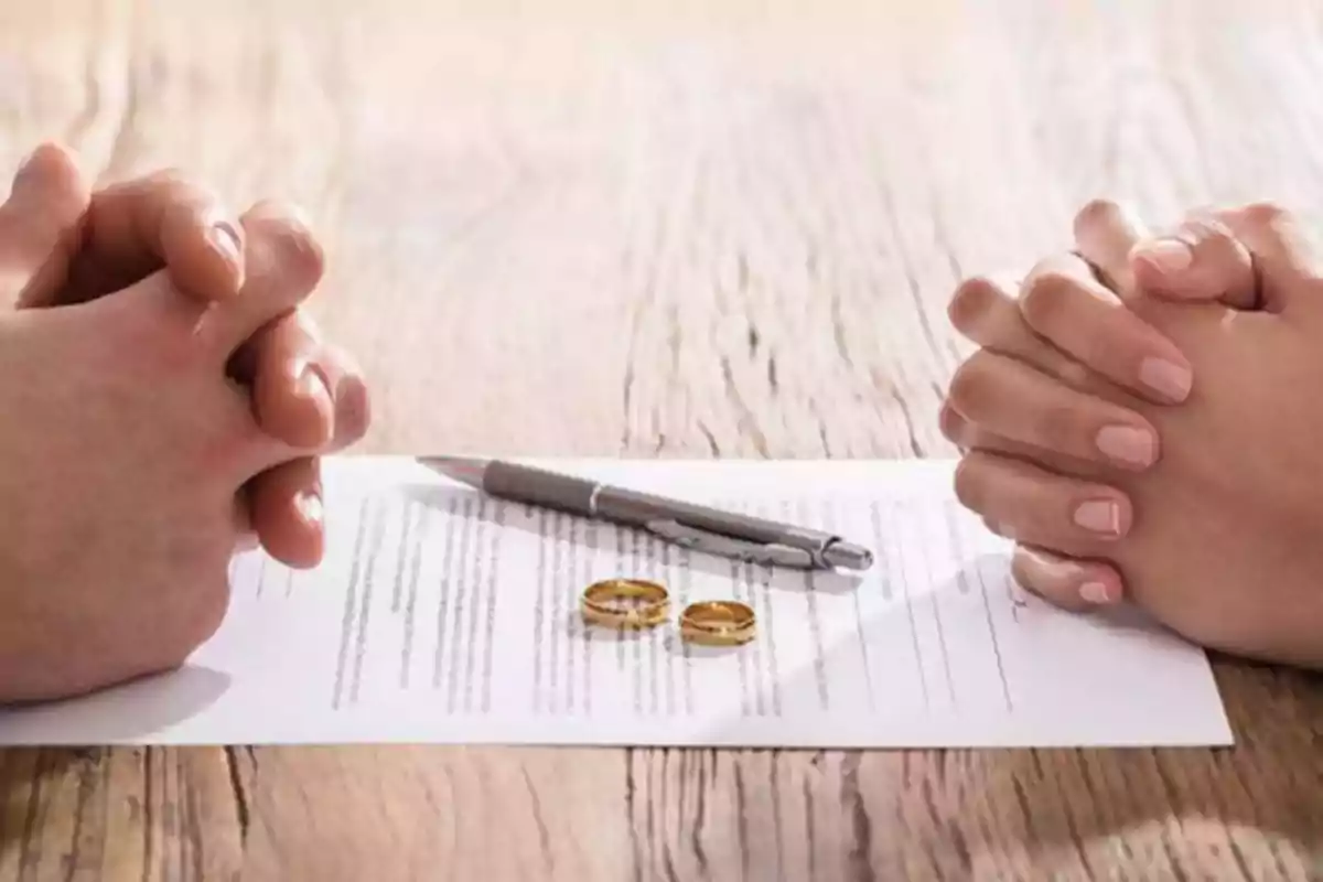 Dos personas con las manos entrelazadas frente a un documento con un bolígrafo y dos anillos sobre una mesa de madera.