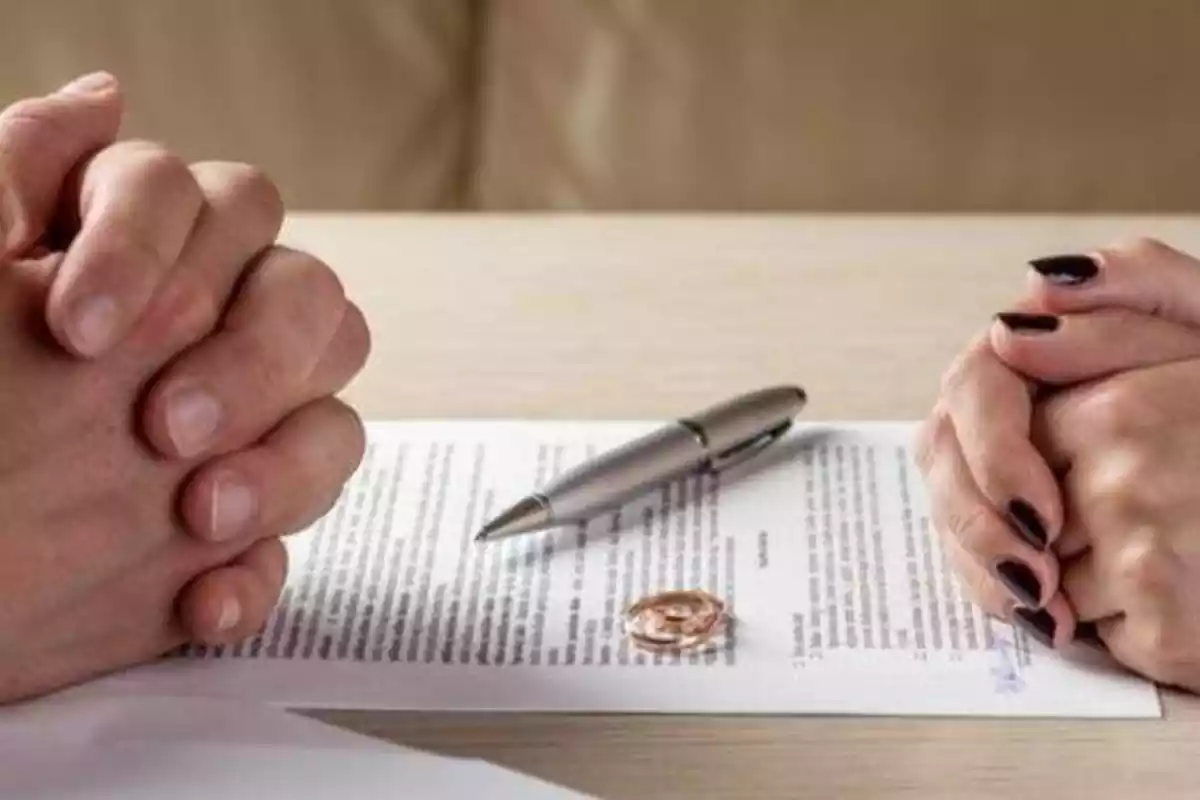 Las manos de un hombre y una mujer con las uñas pintadas enfrentadas. En medio, un papel de divorcio con los anillos encima y un bolígrafo.