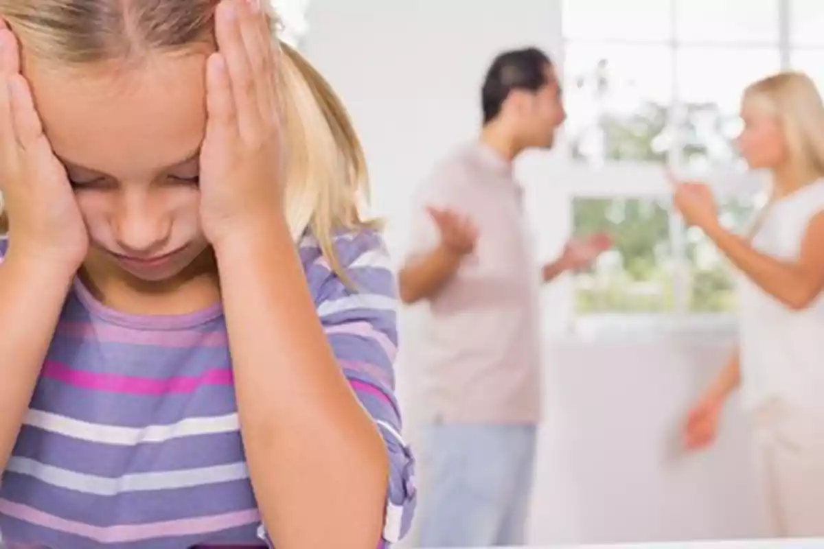 Una niña con las manos en la cabeza parece angustiada mientras dos adultos discuten en el fondo.