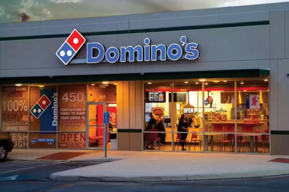 Fachada de una pizzería con el logo de Domino's iluminada al anochecer. Fachada de una pizzería con el logo de Domino's iluminada al anochecer.