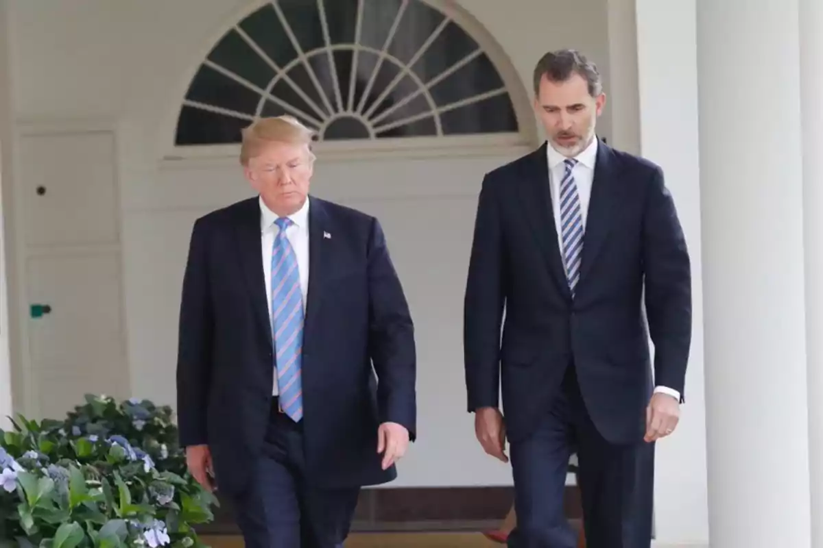 Dos hombres caminando juntos en un pasillo exterior con columnas y plantas.