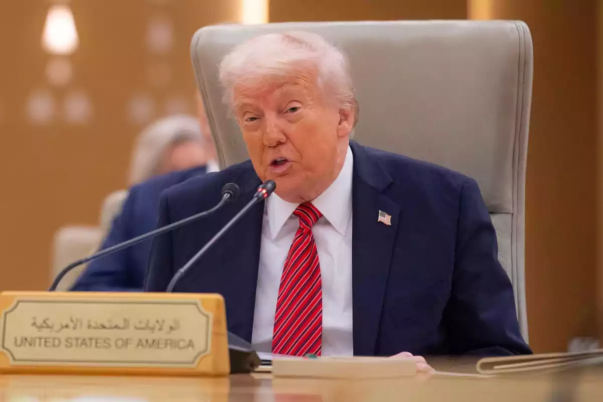 Donald Trump de traje oscuro y corbata roja habla en un micrófono durante una conferencia, con un letrero que dice Donald Trump de traje oscuro y corbata roja habla en un micrófono durante una conferencia, con un letrero que dice