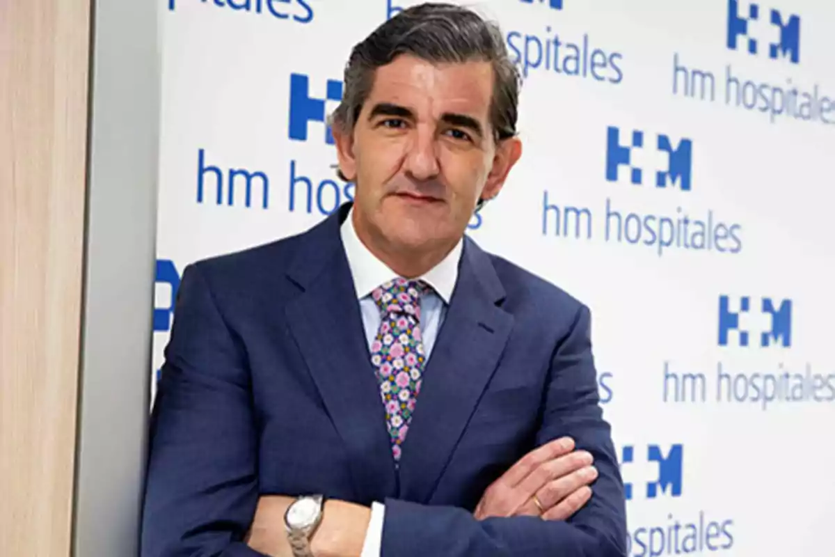 Hombre de traje azul con corbata de flores posando frente a un fondo con el logo de HM Hospitales.