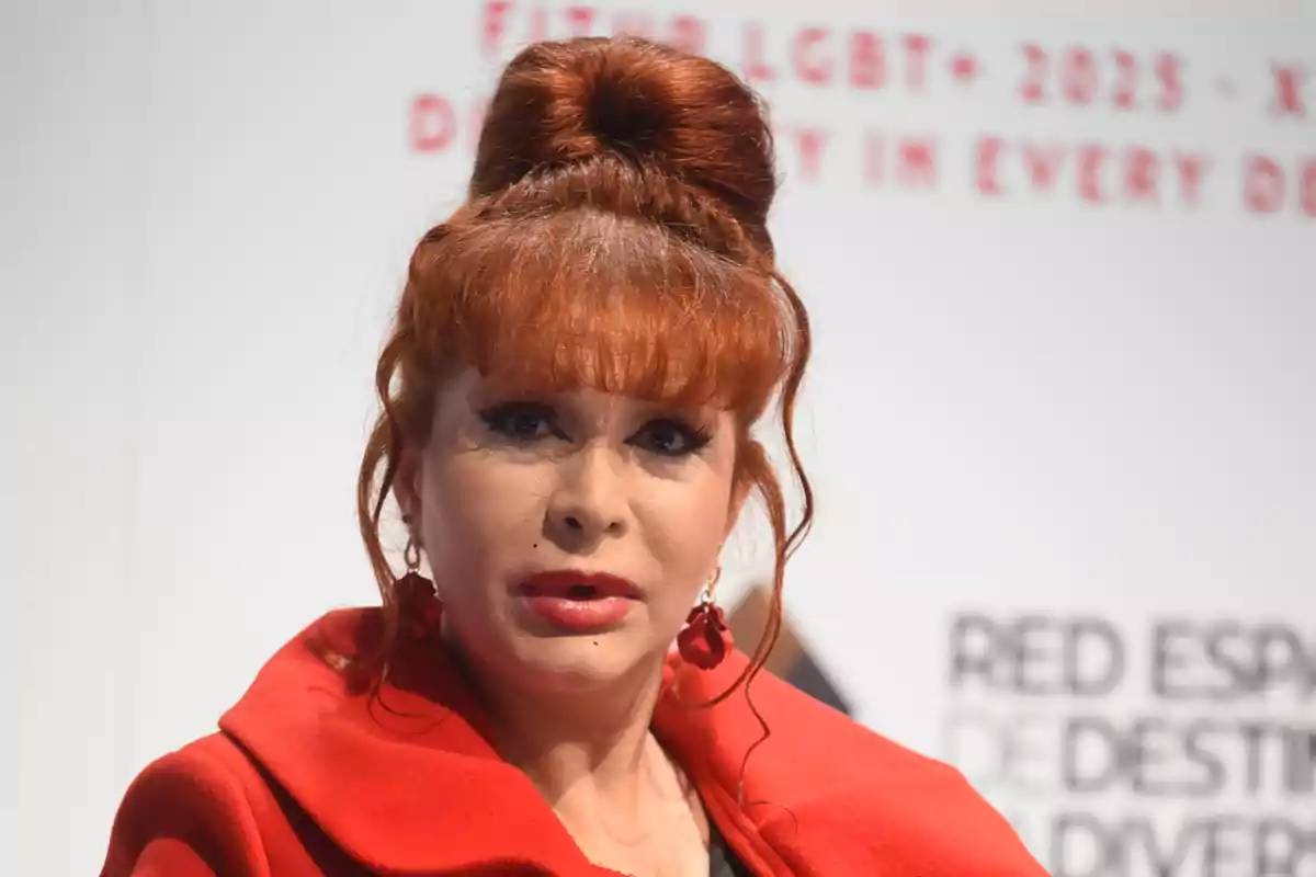 Una persona con cabello rojo recogido y un abrigo rojo, en un evento con fondo de texto borroso.