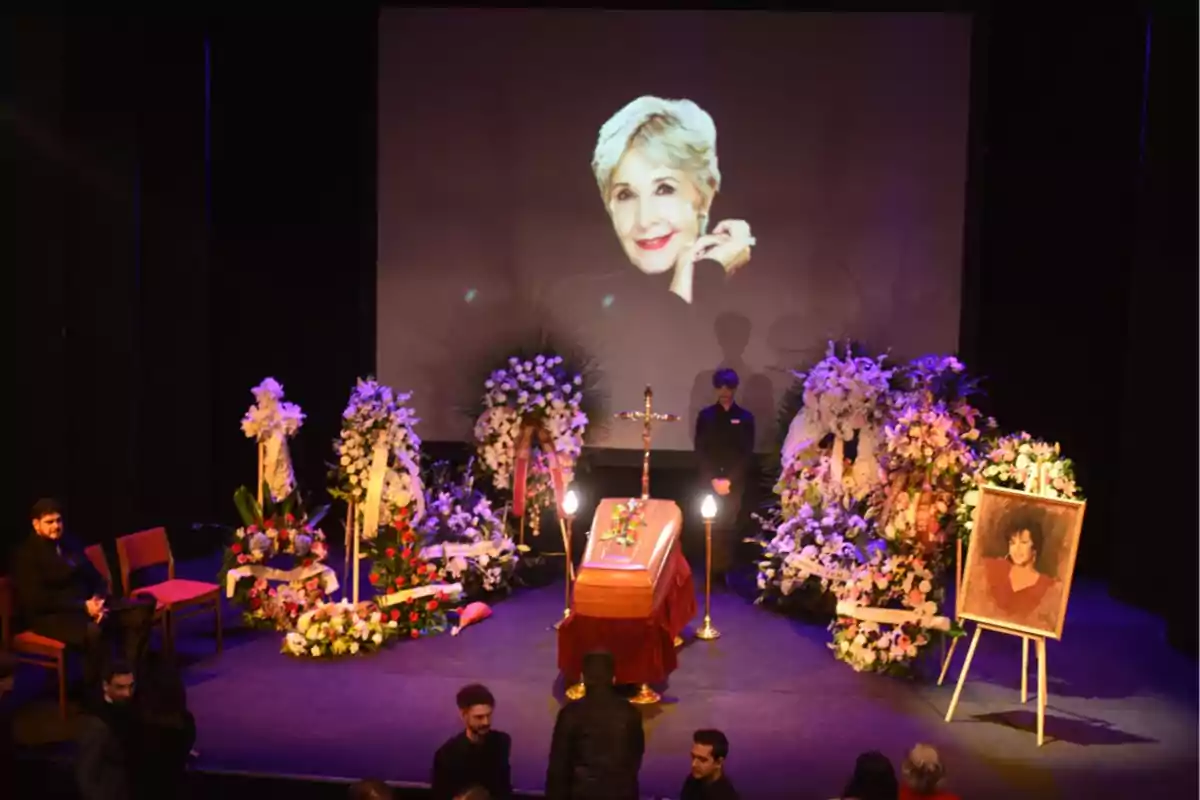 Una ceremonia fúnebre con un ataúd rodeado de arreglos florales, una cruz, y dos retratos de una mujer, uno en un marco y otro proyectado en una pantalla grande.