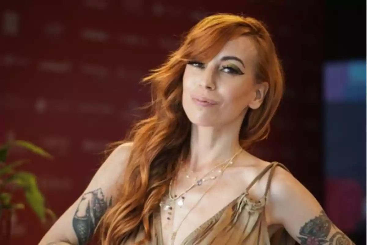 Mujer pelirroja con cabello largo y ondulado, maquillaje llamativo y tatuajes en ambos brazos, posando con una expresión confiada frente a un fondo borroso rojo Mujer pelirroja con cabello largo y ondulado, maquillaje llamativo y tatuajes en ambos brazos, posando con una expresión confiada frente a un fondo borroso rojo