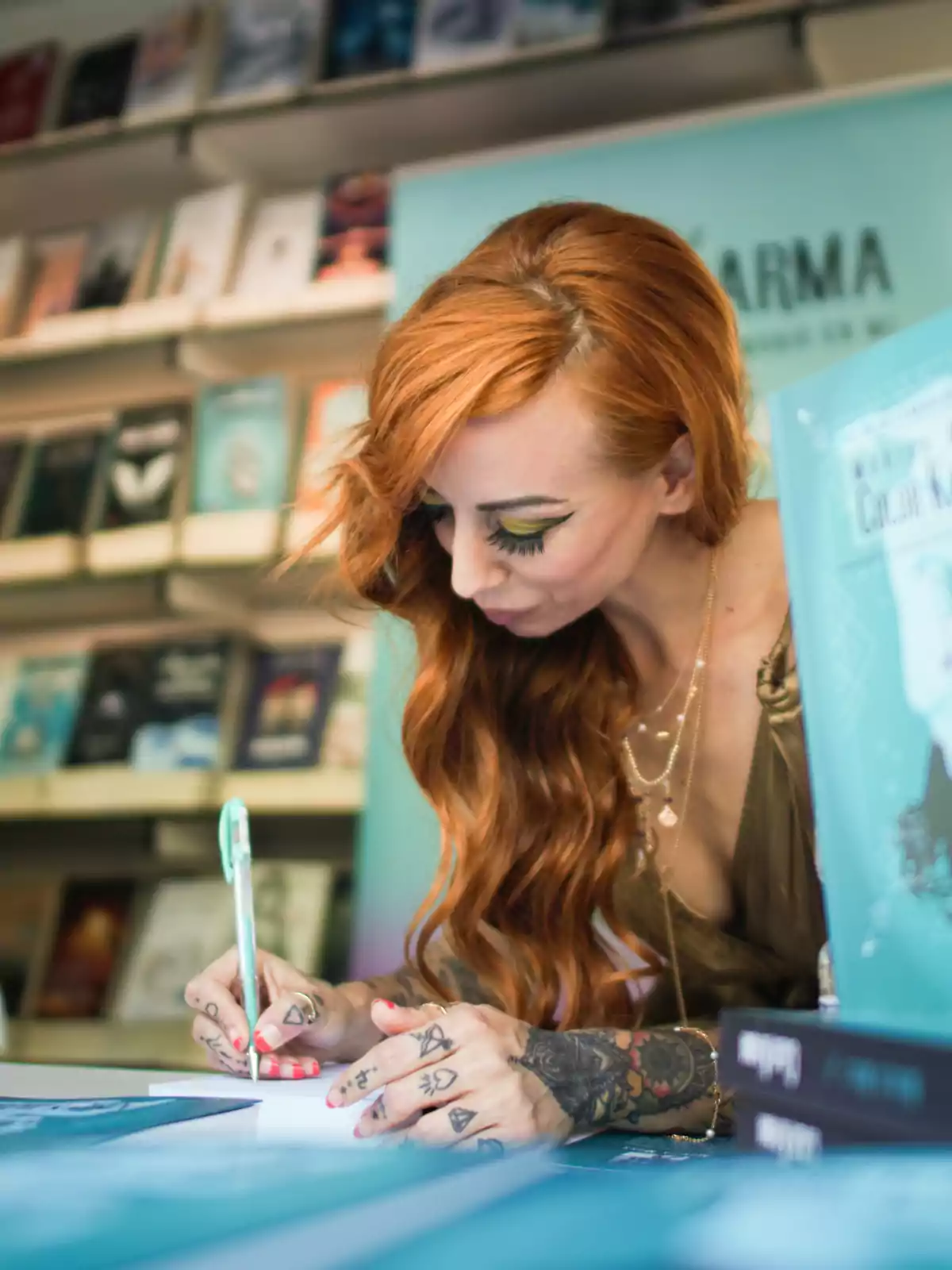Mujer de cabello largo y pelirrojo firmando un libro en una librería rodeada de estantes con libros Mujer de cabello largo y pelirrojo firmando un libro en una librería rodeada de estantes con libros