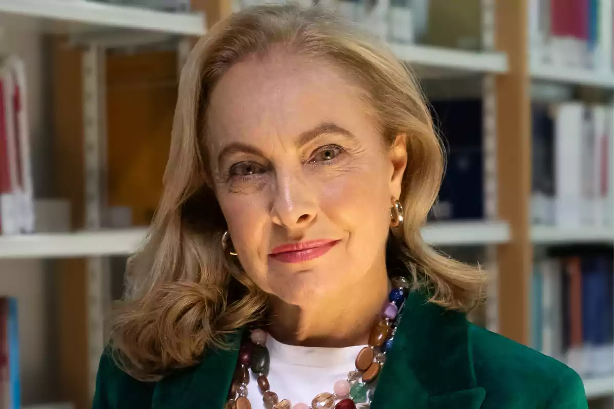 Una mujer de cabello rubio y ondulado, vestida con un blazer verde y un collar de cuentas coloridas, está de pie frente a una estantería de libros.