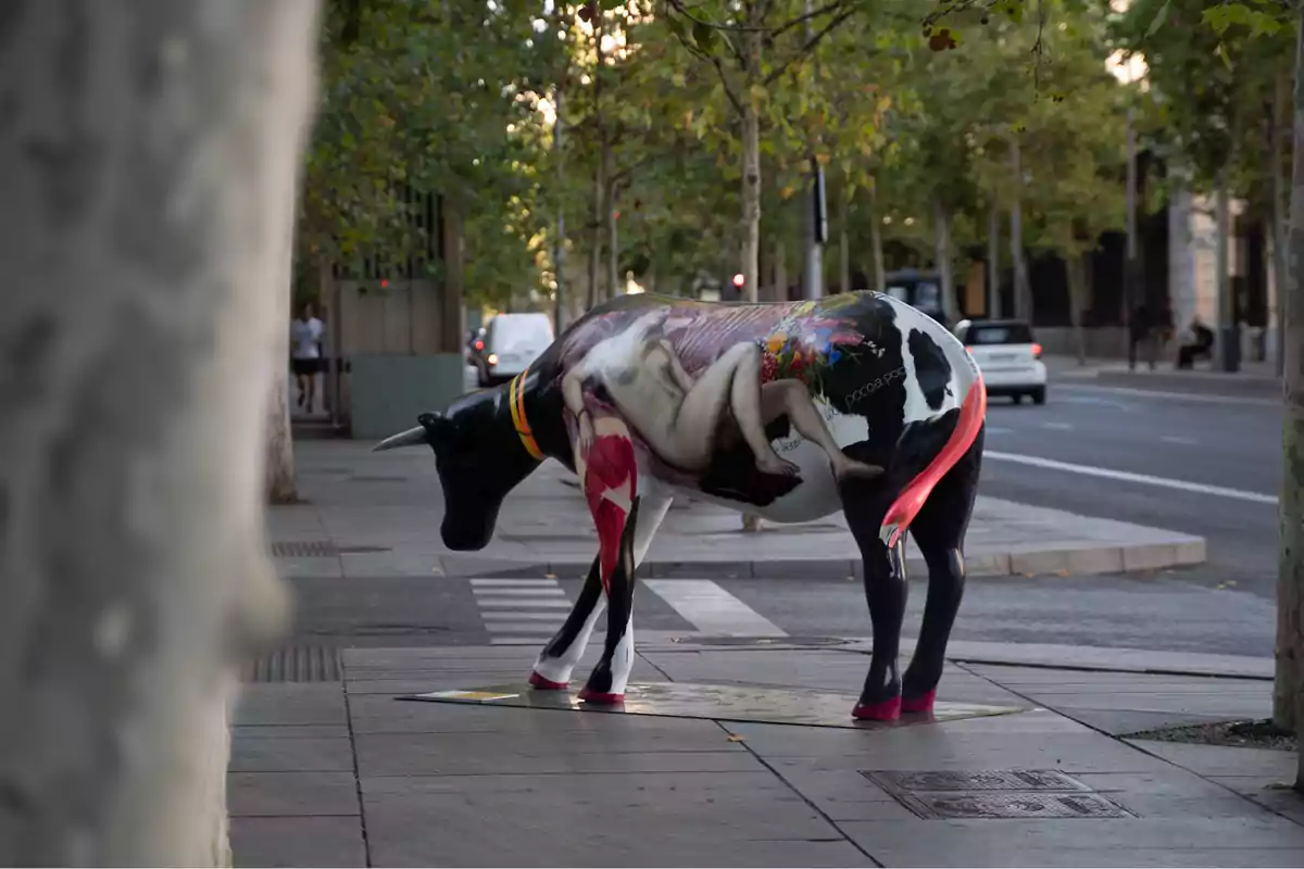 Escultura de una vaca pintada con un diseño artístico en una calle arbolada.