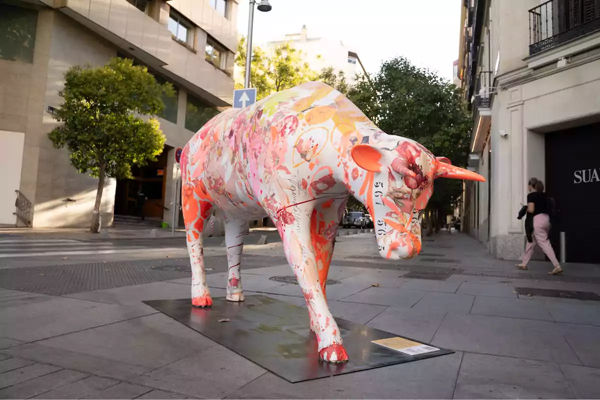 Una escultura de un toro decorado con colores vibrantes y patrones florales se encuentra en una calle urbana, rodeada de edificios y árboles.