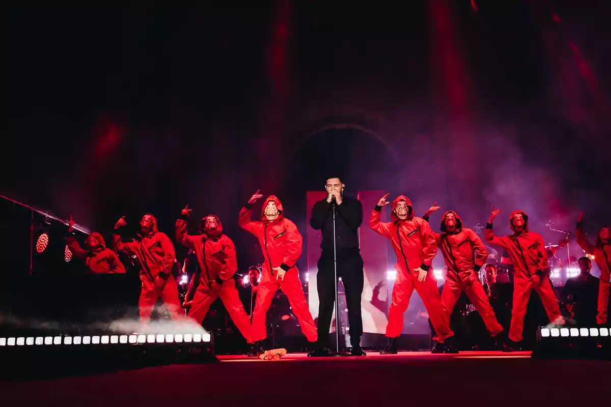 Un cantante vestido de negro se presenta en el escenario acompañado de bailarines con trajes rojos y máscaras, bajo luces rojas y humo teatral Un cantante vestido de negro se presenta en el escenario acompañado de bailarines con trajes rojos y máscaras, bajo luces rojas y humo teatral