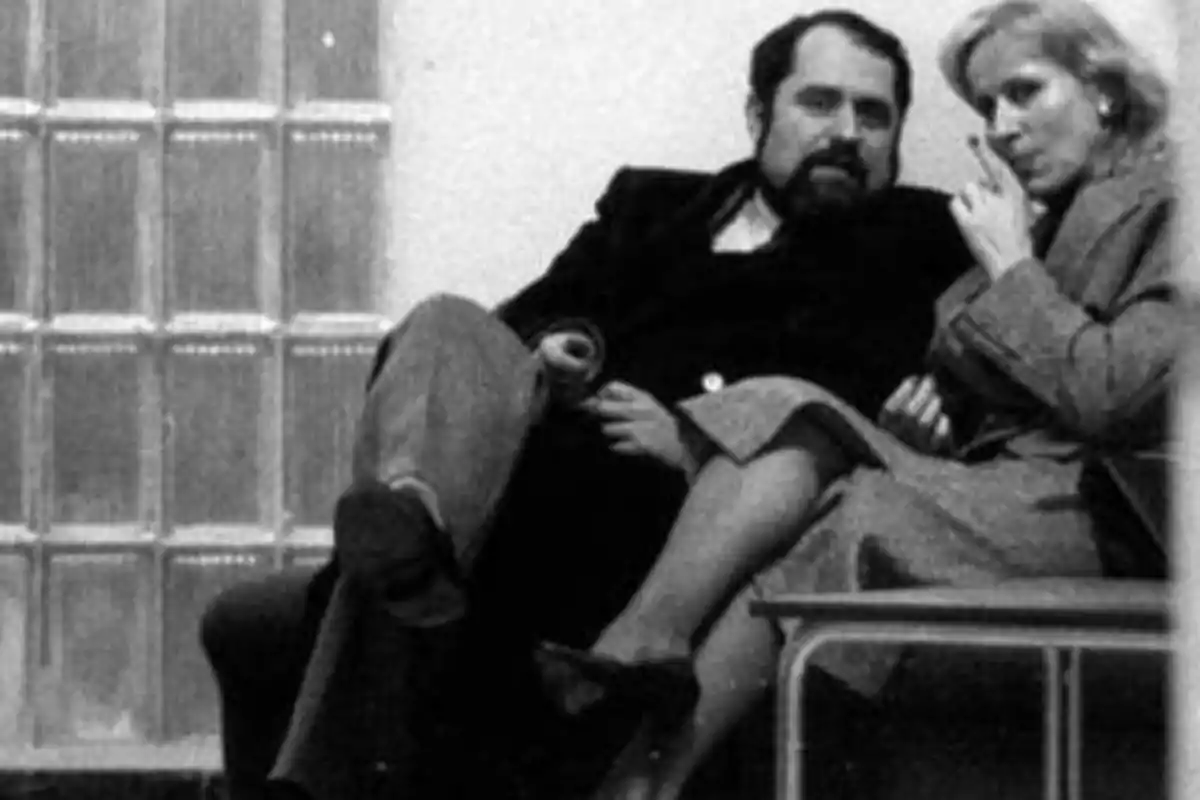 Una pareja sentada en un banco, el hombre con barba y la mujer fumando, ambos con ropa de invierno.
