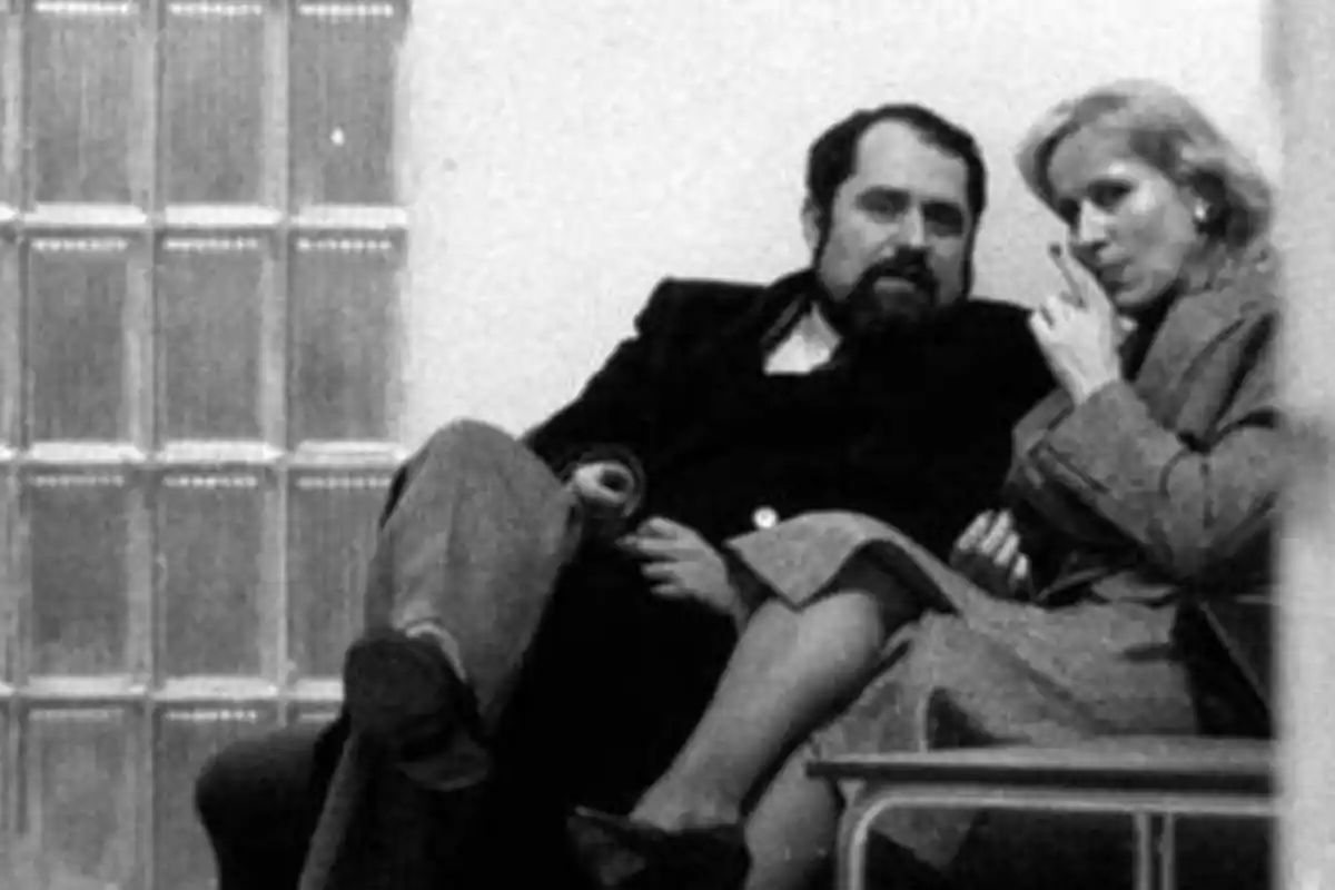 Una pareja sentada en un sofá, el hombre con barba y la mujer rubia, ambos con ropa formal.