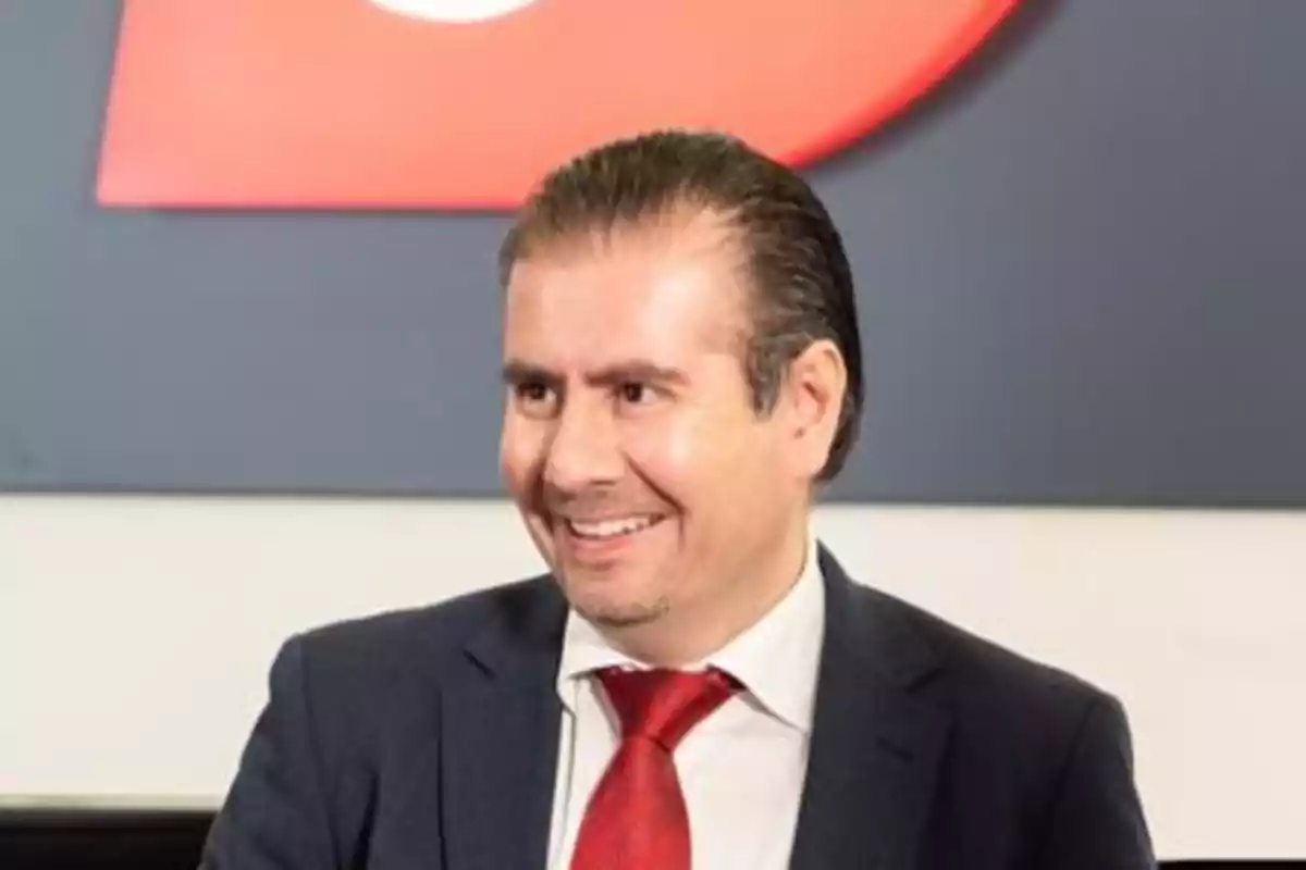 Un hombre sonriente con traje y corbata roja frente a un fondo gris y rojo.