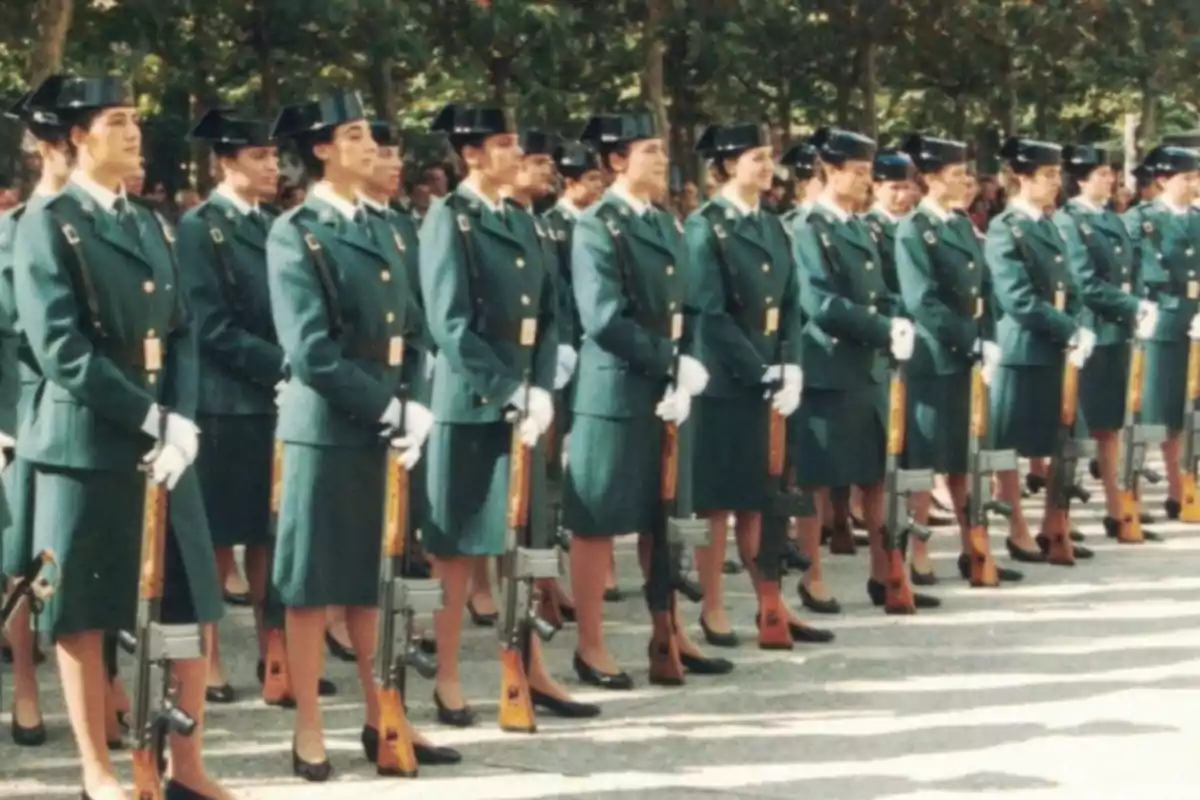 Un grupo de mujeres en uniforme militar verde oscuro, con sombreros y guantes blancos, están formadas en fila sosteniendo rifles.