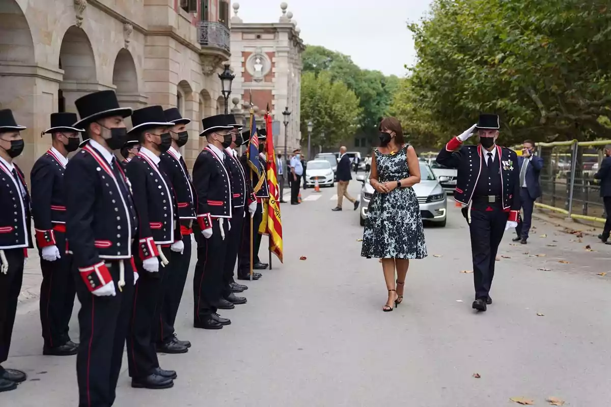 Una mujer camina junto a un oficial que la saluda mientras pasa frente a una fila de guardias uniformados.