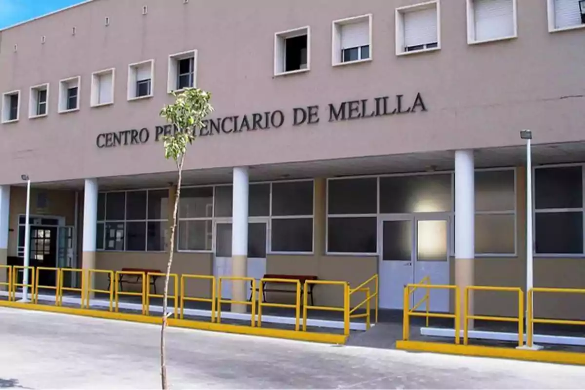 Fachada del Centro Penitenciario de Melilla con barandillas amarillas y un árbol pequeño en primer plano.