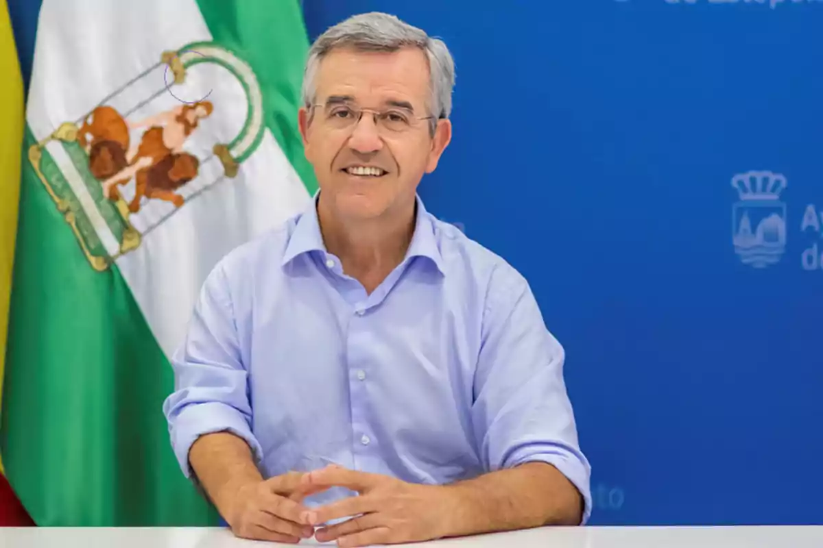 Un hombre con camisa azul claro está sentado frente a una bandera verde y blanca y un fondo azul.