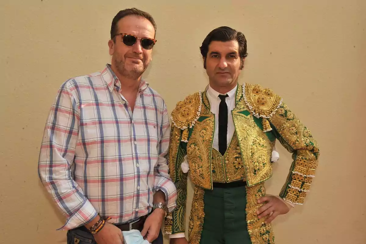 Dos hombres posan juntos, uno lleva una camisa a cuadros y gafas de sol, el otro viste un traje de torero verde y dorado.