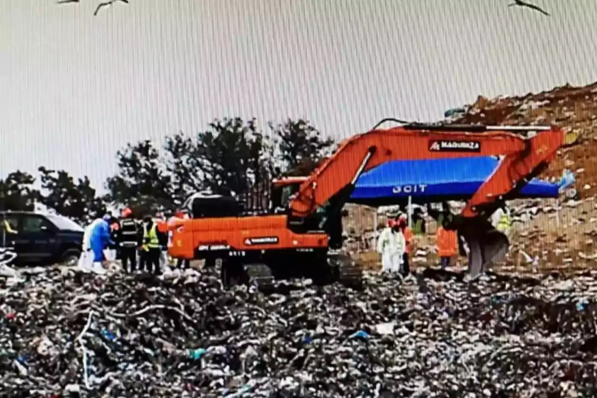 Una excavadora naranja y azul en un vertedero con personas usando equipo de protección.