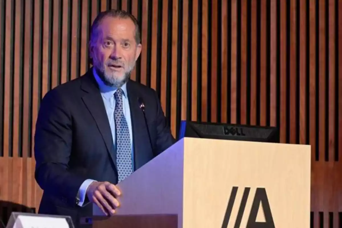 Un hombre de traje y corbata habla desde un podio en un auditorio con fondo de madera. Un hombre de traje y corbata habla desde un podio en un auditorio con fondo de madera.