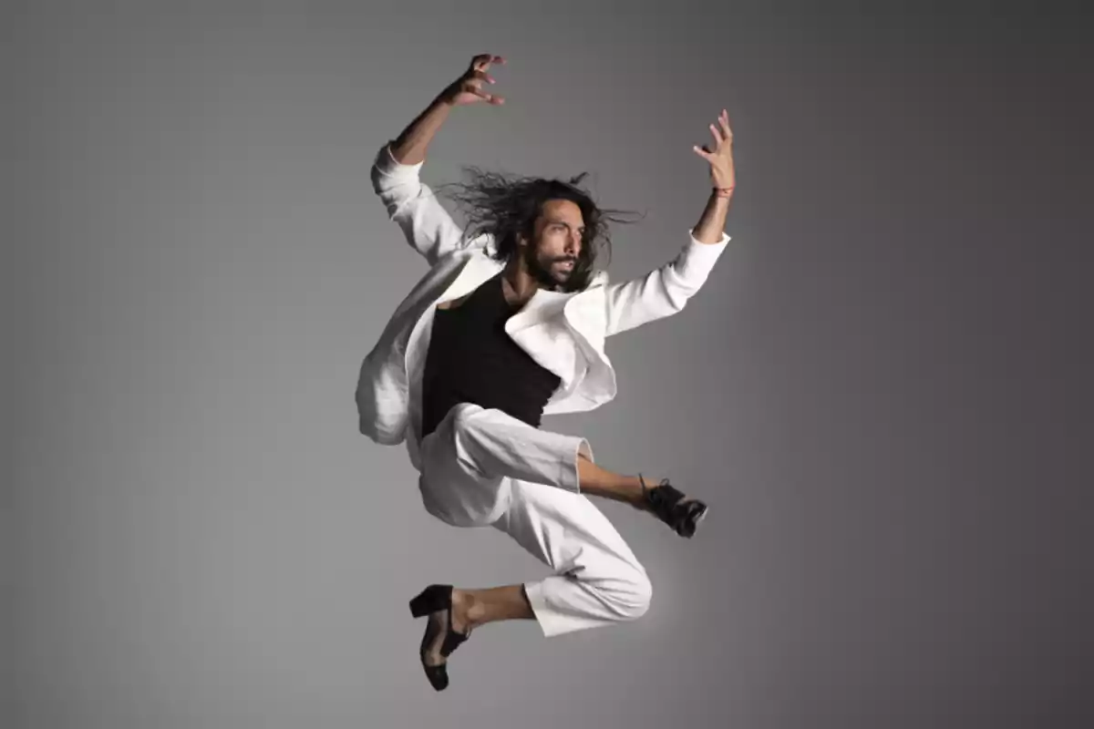 Un hombre con traje blanco y camiseta negra realiza un salto acrobático en el aire.