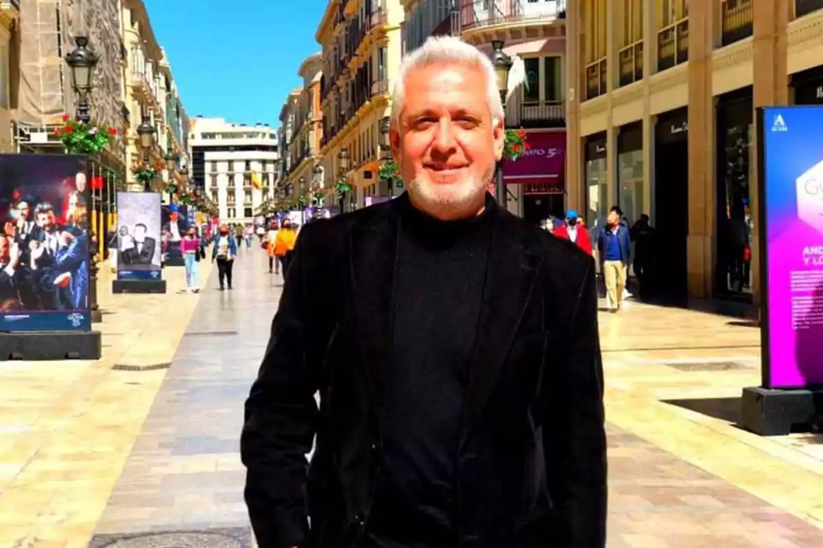 Hombre de cabello canoso y barba, vestido con un suéter negro y chaqueta negra, posando en una calle peatonal con edificios y carteles a los lados, bajo un cielo despejado.