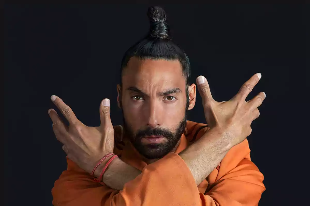 Hombre con cabello recogido y barba en pose con los brazos cruzados frente a un fondo oscuro.