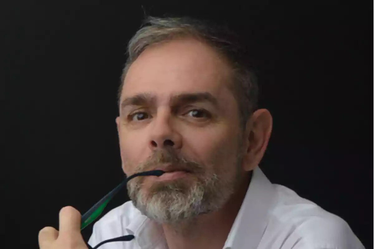 Un hombre con barba y cabello canoso sostiene unas gafas en su boca mientras mira hacia la cámara. Un hombre con barba y cabello canoso sostiene unas gafas en su boca mientras mira hacia la cámara.